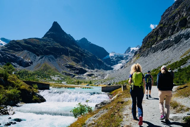 Hikers follow a path beside rushing turquoise water, with rocky peaks and a glacier ahead beneath blue skies.