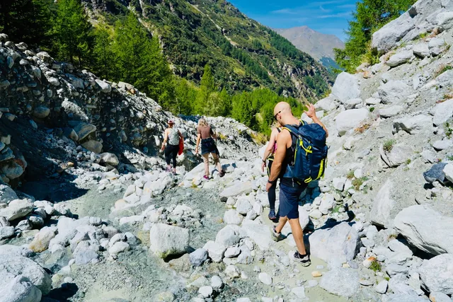 Hikers follow a rocky alpine trail beside a stone retaining wall, surrounded by boulders, trees and steep slopes.