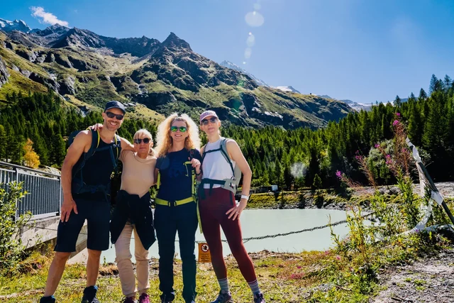 Four hikers stand together beside pale turquoise water, with evergreen forest and rugged mountains beneath a blue sky.