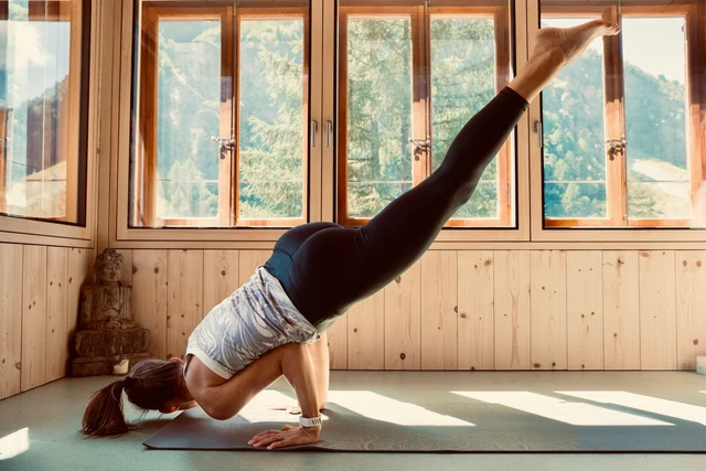 A woman balances on bent arms with her legs extending upward in a sunlit, wood-panelled yoga room.
