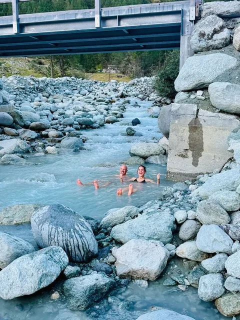 Two smiling bathers give thumbs-up while floating in pale blue river water beneath a bridge, surrounded by large grey boulders.