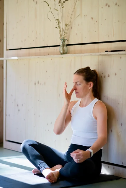 A woman sits cross-legged with eyes closed and one hand beside her nose during breathing practice in a wooden room.