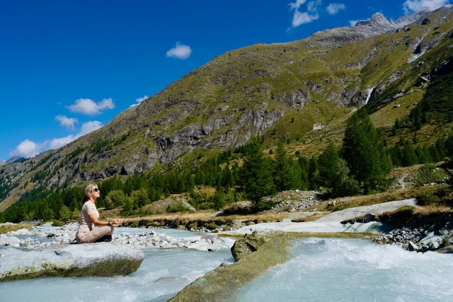 A man sits cross-legged on a rock beside a rushing turquoise river, surrounded by alpine trees and steep mountains.