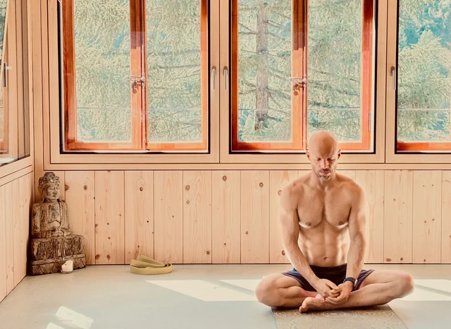 A man sits cross-legged with his hands resting together in his lap in a sunlit wooden yoga room beneath large windows.