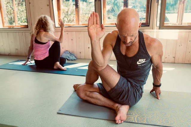 A man twists from a seated yoga posture with one hand raised while a woman practises behind him in a wooden room.