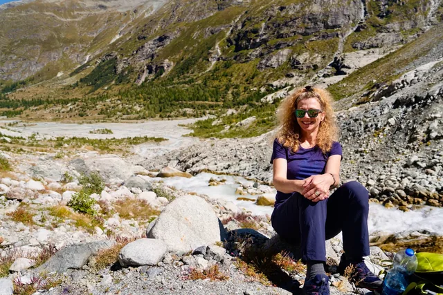 A smiling woman wearing sunglasses rests on rocks beside an alpine river, with a gravel valley and steep mountain slopes behind.
