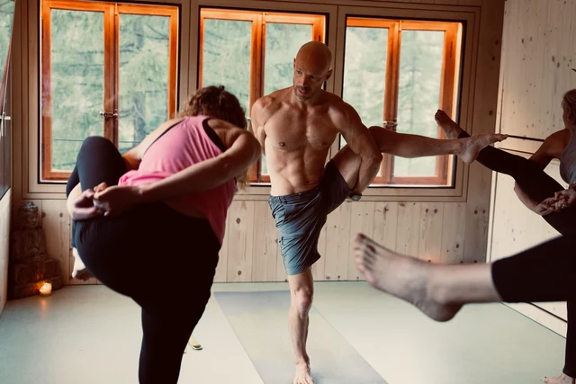 A teacher demonstrates a standing leg balance while participants practise beside him in a wooden yoga room.