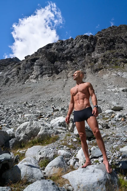 A man in dark swimming shorts stands barefoot on a boulder beneath a rocky alpine slope, looking sideways under a blue sky.