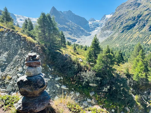 A small stone cairn stands above a forested mountain valley, with rocky peaks and a glacier beneath a clear blue sky.
