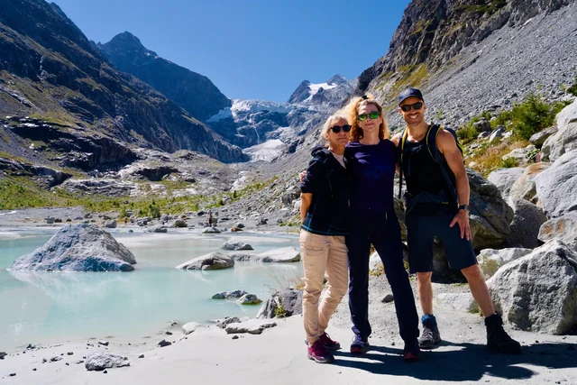 Three hikers smile beside a pale glacial pool, with a glacier descending between steep rocky mountain slopes behind them.