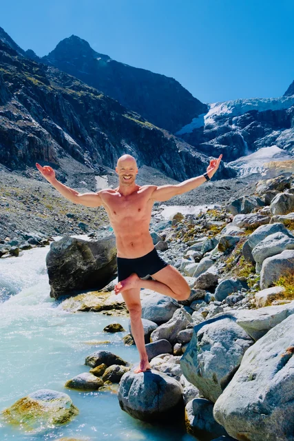 A man balances in Tree pose with both arms raised on a boulder beside a turquoise alpine river, with a glacier behind him.