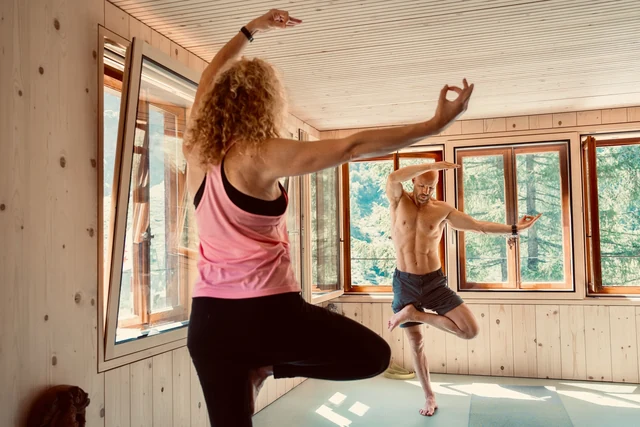 A woman and a man balance in tree pose with arms lifted inside a sunlit wooden yoga room overlooking trees.