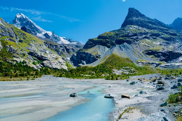 Pale turquoise river channels wind across a broad gravel bed beneath rocky mountains and glacier ice under a bright blue sky.