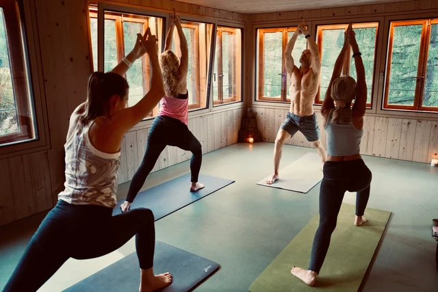 A yoga teacher leads a group in Warrior I with arms raised overhead in a sunlit, wood-panelled room.