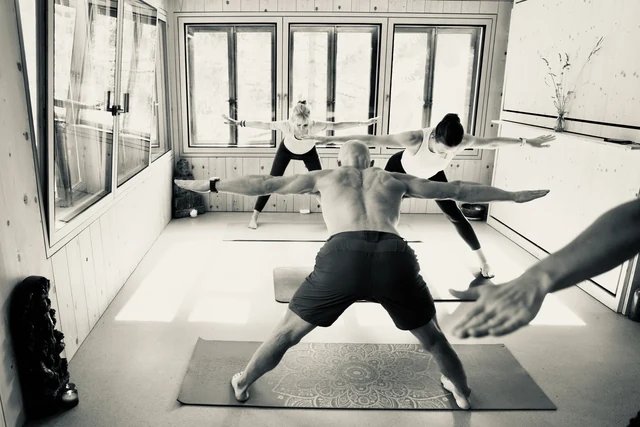 A black-and-white view of a yoga class holding Warrior II with arms extended in a window-lined wooden room.