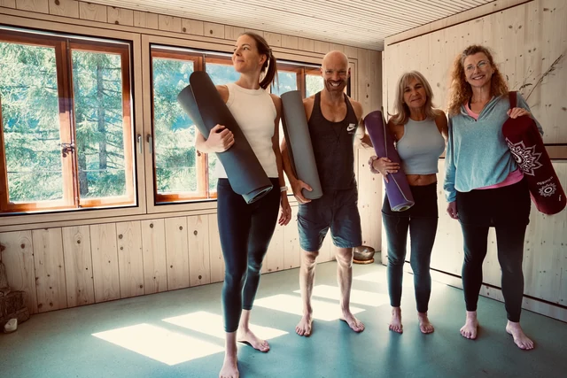 Four smiling people stand together holding rolled yoga mats in a sunlit wooden room with large windows.